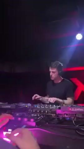 The video features a DJ performing in what appears to be a club setting. The DJ is a young man with dark hair, wearing a dark t-shirt and headphones. He is standing behind a DJ mixing console with two turntables on either side. He is actively manipulating the equipment, mixing music, and paying close attention to the sound. The console is illuminated with bright neon pink and purple lights. In the background, there is a dark wall with an "X" logo in red neon.  A white spotlight is shining down from above. Another person's hands are visible at the very bottom edge of the frame, also lit in pink. The overall environment suggests a dark, lively, and energetic nightclub atmosphere.