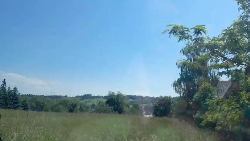 A video taken from a moving vehicle shows a grassy field with a fence on the right side. In the distance, there are trees and hills under a clear blue sky. A white pole with red markings is visible near the fence. The video is approximately 3 seconds long and appears to be recorded during the daytime.