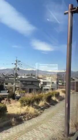 The video shows a view from a moving train or bus, looking out the window at a suburban landscape. There are houses, trees, and power lines visible. In the distance, there are mountains with snow-capped peaks. The sky is blue with some clouds. A child's voice can be heard saying "I think that's in Russia." The camera is moving, so the scenery is constantly changing.