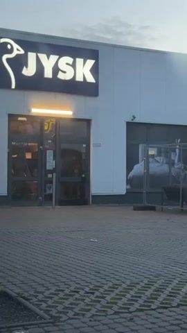 The video shows the exterior of a JYSK store. A car is parked in the foreground, partially obscuring the view. In the window of the store, there is a large advertisement featuring a man and a woman on a sofa. There is also some construction fencing in front of the window. The sky is overcast.