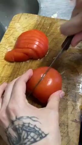 A person is slicing a tomato on a wooden cutting board with a knife. The person's left hand holds the tomato while the right hand wields the knife to create even slices. A stack of sliced tomatoes is already on the board, next to the tomato being sliced. The person has a tattoo on their left hand and is in a kitchen setting.