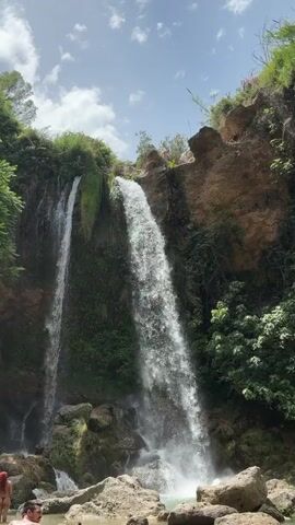 The video shows a waterfall cascading down a rocky cliff face covered in lush green vegetation. The sky above is partly cloudy with a bright sun. In the foreground, there are large rocks and a pool of water where two people are visible. One person is partially submerged in the water, and another is seated on the rocks. Further up the cliff, near the top of the waterfall, a person can be seen standing. The overall scene depicts a natural outdoor environment, likely a park or recreational area, with people enjoying the waterfall.