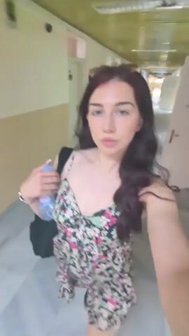 A woman with long, dark hair walks down a narrow hallway. She is wearing a floral print dress and sunglasses on her head. She holds a bottle of water in her left hand. The hallway appears to be indoors and has a textured wall on the right side and a lighter colored wall on the left. The floor is a light marble or tile pattern. The lighting is dim and fluorescent. The woman briefly makes a peace sign with her fingers as she looks into the camera.