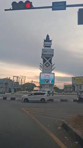 The video shows a street scene with a tall monument in the center, featuring a digital billboard. The sky is overcast, suggesting it might be dawn or dusk. There are traffic lights visible, with a red light illuminated. A blue sign reads "DILARANG MENEROBOS" which translates to "DO NOT BREAK THROUGH". Various vehicles, including a white car and several motorcycles, are present on the road. Some people are riding the motorcycles. The monument has "BSI" prominently displayed on it, along with some text and images on the digital billboard. The environment appears to be an urban or semi-urban setting with buildings and infrastructure in the background. The camera appears to be static, filming the scene from a road perspective.