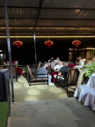 The video captures an outdoor evening gathering at a waterfront venue, likely a restaurant or event space in Tây Nha Trang, Vietnam. The scene is illuminated by decorative red lanterns hanging from a covered ceiling, as well as string lights and ambient lighting, casting a warm glow. A body of water, possibly a river or lake, is visible in the dark background, reflecting distant lights.

Numerous individuals, primarily adults, are present. Initially, several men are shown seated at tables, engaged in conversation. As the camera pans, more groups of people are visible at other tables. The video then reveals an area set up with multiple rows of white, fabric-covered chairs, arranged as if for an audience. People are seen socializing, sitting at tables, standing, and moving around the venue. The atmosphere appears relaxed and convivial. No visible text is present in the video.