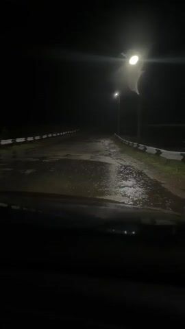 The video shows a dark, wet road at night, seen from the perspective of someone in a vehicle. Streetlights illuminate the road, creating reflections on the wet surface. The road appears to be a bridge or elevated roadway, with guardrails visible on either side. The overall atmosphere is dark and somewhat eerie.