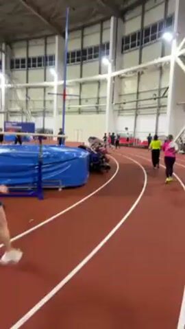 The video shows a group of people running on an indoor track. The track is red with white lines indicating lanes. There are several runners, dressed in various athletic outfits, moving in the same direction along the track.  The surrounding environment is an indoor sports facility with high ceilings, windows along the walls, and bright overhead lighting. A pole vaulting area is also visible on the side of the track, featuring blue mats and equipment. The camera is positioned from the perspective of a runner, giving a first-person view of the race or practice.