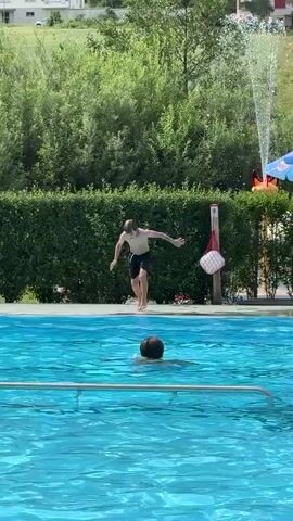 The video shows a person jumping into a swimming pool. They perform a forward somersault and enter the water with their body parallel to the surface. Another person is in the pool, swimming. The background consists of lush green trees and buildings. A water fountain is visible on the right side of the frame. A red and white object hangs from a pole near a white bucket.