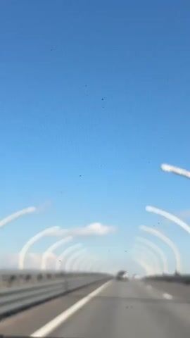 The video shows a view from the front of a car driving along a highway on a sunny day. The sky is blue, with a few white clouds visible. The road is gray and has white lane markings.  The road is lined with tall white arched streetlights. There are other cars on the road moving in the same direction.