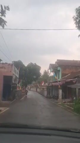The video shows a view from inside a vehicle, looking out at a wet, paved road in what appears to be a rural or suburban area. The sky is overcast and grey. On either side of the road are buildings, some with houses and some with businesses. Indonesian flags are prominently displayed on flagpoles in front of many of the buildings. There are trees lining the road, including a large, leafy tree on the left. A person wearing a dark shirt and pants is walking on the left side of the road. Another person is visible on a motorcycle further down the road on the left. The road has some cracks and potholes. The overall impression is of a quiet, overcast day in a community decorated for a national holiday.