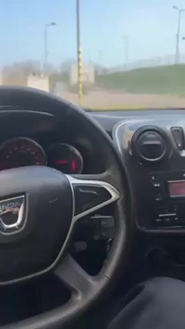 A person is in the driver's seat of a Dacia car. The dashboard and steering wheel are visible, as well as a view out of the front windshield. The radio is on, playing music. The environment visible through the windshield includes streetlights and a grassy area. The day appears sunny.