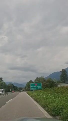 The video shows a forward-facing view from a vehicle driving on a highway. The sky is overcast with gray clouds. On the right side of the road, there is a green road sign indicating distances to two towns: Brissago 20 km and Locarno 9 km. The road has a white line on the right edge and a guardrail. There are trees and green vegetation lining the side of the road. In the distance, mountains are visible. Other vehicles, including a truck, are also seen on the highway. The video is a continuous shot without any notable actions or interactions.