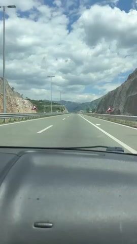 The view from inside a vehicle looking out through the windshield onto a straight, well-maintained highway. The road is divided into lanes by white lines, and there are green road signs on the side of the road, as well as triangular signs with downward-facing arrows indicating a slope. The sky is blue with scattered white clouds. Mountainous terrain flanks both sides of the highway. The interior of the car, including part of the dashboard, is visible in the foreground.