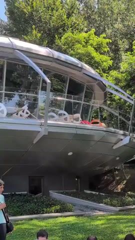 The video shows a futuristic-looking building with a curved, metallic exterior and large glass windows. The building is situated amidst lush green trees and foliage, suggesting a natural or park-like setting. There is a balcony or deck area on the upper level of the building, accessible through the glass windows. On this deck, several white chairs and some colorful cushions (red and orange) are visible, implying a space for relaxation or outdoor seating. Below the building, a grassy area is visible, and in the foreground, several people's heads and shoulders can be seen, indicating that the viewer is likely standing on the ground level, looking up at the structure. The overall impression is one of modern architecture integrated with nature.
