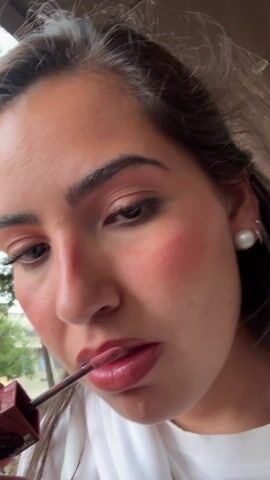 A close-up shot shows a young woman applying lip gloss. She has light skin, brown hair pulled back, and is wearing pearl earrings. The woman is focused on applying the lip gloss, looking down at her lips with a slight smile. She is wearing visible makeup, including eye shadow and blush. The background is slightly blurred and shows greenery and a building in the distance. The overall lighting suggests an outdoor setting during the daytime.