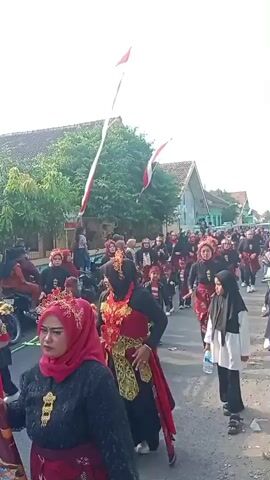 A group of people, many wearing traditional red and black clothing with headscarves, are marching down a street. Some are carrying what appears to be a makeshift stretcher or platform made of green material and wooden poles. There are children in the procession as well, also in traditional attire. Two red and white flags on long poles are visible, one held by someone in the group and another extending high into the sky. The background shows houses with tiled roofs and trees lining the street. The weather appears to be daytime and overcast.