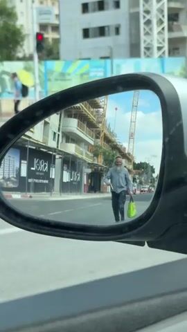 A rear-view side mirror shows a man standing in the middle of the street. The man has a long, gray beard and is wearing a dark shirt and pants. He is holding a neon green shopping bag in one hand and looking at a cell phone in the other. In the background, there are multiple cars driving on the road, buildings under construction, and utility poles. A white truck passes by the car taking the video.  The car taking the video is stationary.