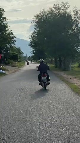 A rear view of a person riding a motorcycle down a paved road. The person is wearing a dark shirt and a helmet. The motorcycle is red and silver and has a license plate with the numbers 86-H6-12. There are other people on motorcycles in the distance. The road is lined with trees and grass. In the background, there are mountains and a cloudy sky. The video appears to have been filmed from another motorcycle following behind.