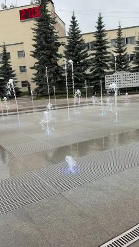 The video captures a public square with a modern fountain. Several jets of water shoot upwards from the ground, some illuminated with blue light. The square is paved with stone tiles and metal grates. In the background, a building with a large red digital clock displaying 27:08 is visible, along with several tall evergreen trees. Further back, more people are seen interacting with the fountain and walking through the square. The environment appears to be an urban setting with residential and commercial buildings surrounding the plaza.