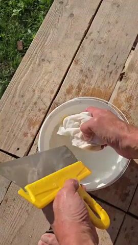 A person is cleaning a tool with a yellow handle and a metal blade in a bucket of water. They are using a white cloth to wipe the blade and a hand to help clean the bucket. The person is standing on a wooden surface outdoors.