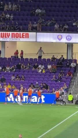 The video shows a soccer field with players on it, wearing orange vests over purple uniforms. Spectators are seated in purple bleachers behind them. A coach or staff member, wearing a black jacket with white stripes, is talking to one of the players. A security guard in a yellow vest is also visible. The field is green, and there are blue and white advertisements along the sideline. The overall atmosphere appears to be a professional sporting event.