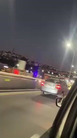 The video, shot from inside a moving car at night, shows a view of an urban landscape. Streetlights illuminate the roadway, and traffic is visible, including a silver car with a rack on top and a yellow taxi. The city's skyline is visible in the background. Loud wind noise is present.