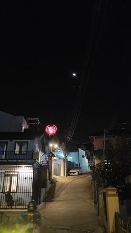 The video depicts a narrow, gently uphill cobblestone street at night, illuminated by streetlights and the soft glow emanating from a window in a house on the left. The houses lining the street are painted in light blue and white tones. A car is parked near the end of the street. A red star-shaped light fixture is prominent atop a utility pole. Power lines traverse overhead. The night sky is dark, and a crescent moon is visible. A motorcycle is parked behind a black metal gate to the left of the frame. A figure walks by the car parked at the end of the street.