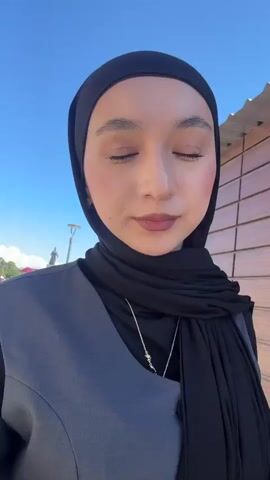A young woman wearing a black hijab, gray vest, and a gold necklace is standing outdoors on a sunny day. She is looking directly at the camera. Behind her, there is a building with a brick wall, a street, and signs in a foreign language. The background includes a bright blue sky. The camera seems to be hand-held, creating slight movements.