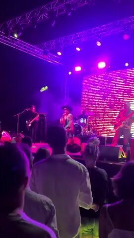 The video shows a live concert performance. In the foreground, the backs of several concertgoers are visible, facing the stage. On the stage, a female singer is performing center stage, wearing a hat, a brightly colored patterned jacket, and jeans. She is holding a microphone and appears to be singing. To her left, a male guitarist is playing an electric guitar. To her right, a drummer is seated behind a drum kit. Behind the performers, a large screen displays a pink and purple patterned graphic. The stage is lit with purple and pink stage lighting, creating a vibrant atmosphere. Smoke or haze is present on the stage, enhancing the lighting effects. The overall scene depicts a dynamic and energetic live music event.