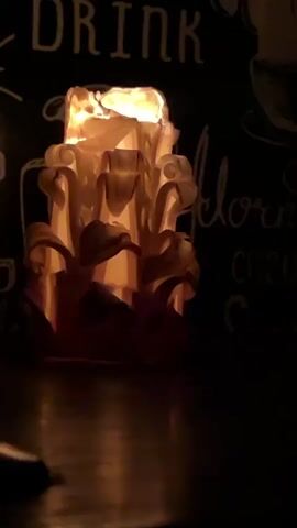 A close-up, low-angle shot of a carved candle burning in the dark. The candle is ornate, with layers of wax carved into petal-like shapes. The flame at the top is bright and illuminates the intricate details of the candle. In the background, out of focus, there are chalk drawings on a dark surface, with the word "DRINK" visible at the top. The lighting is dim and atmospheric, with reflections visible on the surface below the candle. The video is short, with the timestamp indicating it lasts only a few seconds. There are no people or animals visible in the frame.
