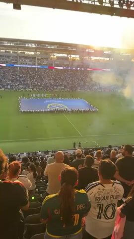 This is a vertical, full-shot video taken from the stands of a soccer stadium. In the foreground, a crowd of spectators are visible from behind, seated in dark gray stadium seating. Some are wearing jerseys, one of which has the name "RIQUI PUIG" and the number "10" on the back. In the mid-ground, a large group of players are standing on the green soccer field holding a blue banner with a gold logo. Another group of people are lined up on the field. In the background, more spectators fill the stadium stands, and a large American flag is displayed on a screen above them. The sky is bright with a golden sunset, and plumes of white and orange smoke are rising from the field, creating a dramatic effect. The overall atmosphere appears to be that of a pre-game ceremony.