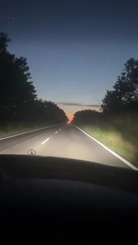 The video shows a first-person perspective from inside a vehicle traveling on a road at dusk. The road is paved and has white lane markings. Trees line both sides of the road. The sky is a deep blue with hints of orange and pink near the horizon, indicating sunset. The vehicle's headlights are on, illuminating the road ahead. The Mercedes-Benz logo is visible on the hood of the car. There are no people visible in the video. The overall mood is serene and tranquil.