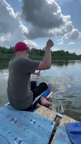 A person wearing a red cap and a grey t-shirt is sitting on a makeshift raft on a body of water. The raft appears to be constructed from inflatable tubes and wooden planks. The person is holding a paddle and is in the process of rowing. The water is calm, with ripples visible around the raft. The background consists of a treeline and a sky with large, white clouds. The perspective is from behind and slightly to the side of the person. The raft has a blue covering with white anchor and ship wheel patterns.