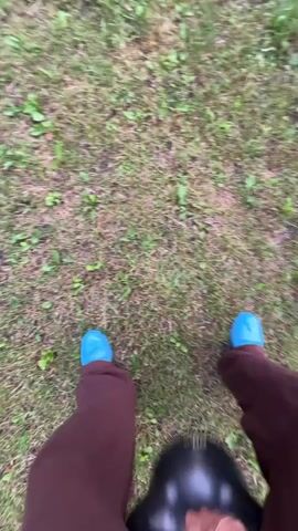 The video shows a first-person perspective of someone walking on grass. The person is wearing blue shoes and brown pants. In the video, the person throws a black object to the ground, then continues walking. The object appears to be a helmet. The environment is outdoors with grass and some greenery visible.