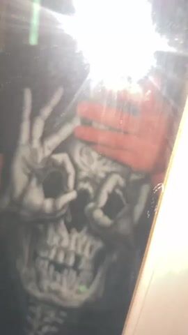 The video shows a person taking a selfie in a mirror. The person is wearing a black t-shirt with a graphic design of a skull with hands covering its eyes. The skull design is monochromatic and detailed. The person's hand holding the phone is visible in the top right corner of the frame, and a bright flash from the phone's camera illuminates the scene. The background appears to be an indoor setting, possibly a room with white trim around the mirror or doorway. The person is wearing red shorts, which are partially visible at the bottom of the frame. The video is very short, lasting only a few seconds, and is characterized by quick movements and the glare of the camera flash.
