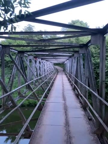 The video shows a slow panning shot of a metal truss bridge spanning a body of water. The bridge has a narrow walkway with two metal tracks. Green trees and foliage are visible on either side of the bridge, and the sky is overcast. In the distance, on the left side, a brick building can be seen through the trees. The camera movement is smooth, suggesting it's mounted on a stable surface or being held by a person walking slowly. There are no people or animals visible in the frame. The overall atmosphere is quiet and natural.