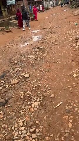 The video shows a dirt road with puddles and stones. There are several people walking on the road, some wearing traditional clothing. There are also buildings on either side of the road, some of which appear to be shops. The sky is overcast. The video pans to the left and then to the right.