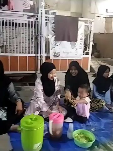 A group of people, including women and children, are gathered on the ground, sitting on blue and green tarps. They appear to be having a meal, with food and drinks laid out in front of them. The environment looks like an outdoor space, possibly a yard or patio, with structures and lighting visible in the background. Some individuals are wearing headscarves. The lighting suggests it is evening or night.