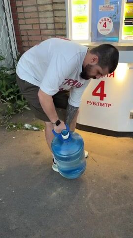 This video shows a man carrying a large, blue, five-gallon water jug with a blue handle. He is wearing a white t-shirt with red lettering, olive green shorts, white socks, and white athletic shoes. He has a beard and a tattoo on his left arm. The man is walking on an asphalt surface, and behind him is a white dispensing machine with a red "4" and "РУБ/ЛИТР" (RUB/LITRE) sign, indicating a price promotion. There's also a brick wall to the left and some greenery. The lighting suggests it's either early morning or late evening. The man appears to be putting in some effort to carry the heavy jug.