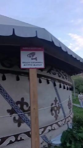 A security camera is visible on a yurt. The yurt is decorated with traditional patterns and tassels. The camera is mounted on a pole and has a red warning sign with a camera icon and text in an unfamiliar language. The video cuts out briefly and then shows a close-up of the yurt's fabric decorations.