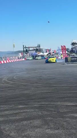 A bright blue and yellow sports car drifts on a black asphalt track, emitting smoke from its tires. The car is angled sharply, with its front tires turned out and its rear tires skidding. In the background, a crowd of spectators is visible behind a white and red barrier. Beyond the crowd, tents and structures suggest an event or festival. The sky is clear and blue, with distant hills visible. The video is shot from a low angle, giving the impression of being close to the action.