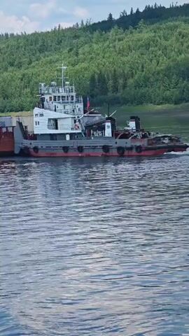 A small tugboat, identified by the text "PT-729" visible on its side, navigates a wide, calm river. The boat is pushing a barge loaded with shipping containers. The tugboat has a white superstructure with multiple windows and a bridge on top, and a grey hull with red accents and black fenders. A Russian flag is visible on the boat's mast.

The river is calm with gentle ripples reflecting the sky. The background is dominated by a lush, green hillside covered in dense forest, suggesting a remote, natural setting. The trees are a mix of deciduous and coniferous species. The sky above is partly cloudy with patches of blue. The overall scene suggests a bright, likely daytime with mild weather.

The activity is the transportation of goods via the river, a common practice in regions with extensive waterways. The presence of the tugboat and barge indicates a working vessel engaged in cargo transport. The environment appears to be a natural, possibly undeveloped area, consistent with the provided location context of Podkamennaya Tunguska, Russia. There are no visible people clearly identifiable on the boat or the barge. The overall mood is one of quiet industrial activity within a serene natural landscape.