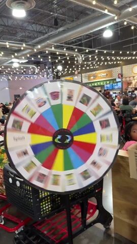 The video shows a colorful spinning wheel at what appears to be a promotional event, probably in a supermarket or food hall. The wheel, mounted on a stand, has sections with different images of food products. The words "SPIN TO WIN" are prominently displayed in the center. In the background, there is a bustling food court with many people sitting at tables eating. There is a table near the spinning wheel that is covered with various snacks, including bags of chips and instant noodles. A person is reaching into a box on the table, pulling out a bag of green chips. The setting is well-lit with string lights overhead, and there are shoppers and employees around, suggesting a busy and active environment.