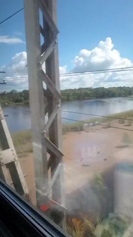 The video shows a view from a moving train crossing a river. The sky is blue with white clouds. The river is wide and dark, with trees lining the far bank. A steel bridge structure is visible in the foreground, with part of the train's interior in the bottom left corner.