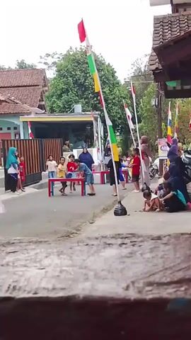 A group of children and adults are gathered on a street, possibly for a local celebration or event. Several flags, including Indonesian flags, are displayed on poles. In the center, a group of children are engaged in a game that appears to involve a table and possibly some sort of competition. Some adults are observing the children, while others are standing or sitting nearby. The environment suggests a residential area with houses and trees in the background.