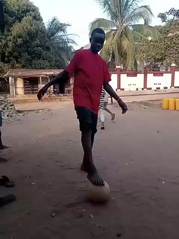 The video shows a young man in a red t-shirt and dark shorts barefoot, bouncing a soccer ball on his foot in a dirt area. Other children are visible in the background, one barefoot, and another in a striped shirt. There are also several sandals on the ground near the camera. In the background, there is a small, rundown building, some trees, a palm tree, and a white wall with a red trim. Yellow plastic barrels are also visible. The light suggests it is daytime.