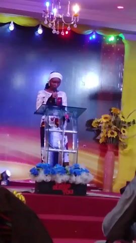 Here is a detailed description of the video:

A woman is standing behind a podium on a red platform, giving a speech. She is wearing a white head covering and a long-sleeved dress with colorful patterns. The podium is clear with white bars. There are flowers in vases on either side of the podium. Behind the woman is a backdrop with an abstract design in blues and oranges. 

There is a chandelier hanging above the stage and colorful lights illuminating the scene. In the foreground, there are people seated in the audience, partially visible at the edges of the frame. A man is seated in front of the camera. 

The camera angle shifts slightly during the video.