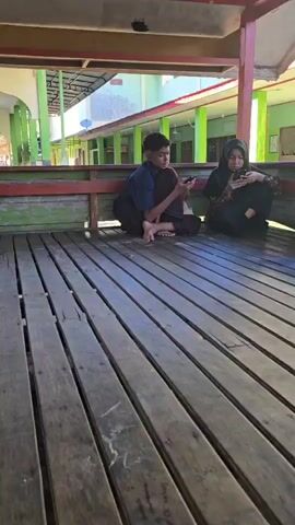 The video shows a group of people, likely teenagers and young adults, sitting on a wooden deck under a covered structure. The structure appears to be open-air, with a red and yellow trim. In the background, a building with green accents and archways is visible, possibly a school or community center. Most of the individuals are wearing headscarves and modest clothing, suggesting a cultural or religious setting. Some are looking at their phones, while others are engaged in conversation, indicating a relaxed social gathering. The wooden planks of the deck are weathered and show signs of age. The overall atmosphere seems casual and informal.