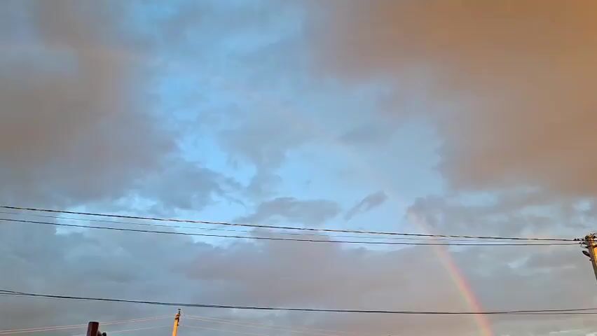 A video clip shows a rainbow in the sky. The sky is filled with clouds, and there are power lines in the foreground. The rainbow is faint and appears to be a double rainbow. The video is shot from a low angle, looking up at the sky.