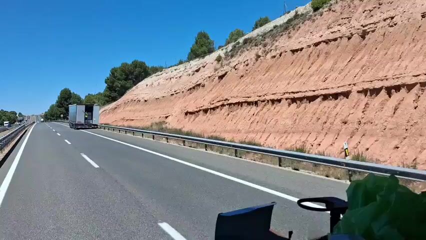 The video shows a highway scene with a large semi-trailer truck in the right lane. A person wearing a yellow safety vest is visible in the open rear door of the truck's trailer. The truck appears to be stopped or moving very slowly, as the camera perspective suggests it is being overtaken by another vehicle (presumably the one recording the video).

The setting is a rural or semi-rural area with a large, reddish-brown rocky embankment on the right side of the highway. The embankment has distinct layers and appears to be a natural geological formation. Pine trees are visible on top of the embankment and along the left side of the highway in the distance. The highway itself is paved with white lane markings and a metal guardrail separates the roadway from the embankment.

The time of day is clearly daytime, with bright sunlight and a clear blue sky, indicating fair weather. The overall impression is a sunny day on a road trip or journey through a somewhat arid landscape.

There is some text visible on the side of the trailer, but it is partially obscured and not clearly legible. Some of the letters may be "MULLER" or similar.

The activity depicted is the operation of a truck on a highway, with a person attending to the trailer. The situation of the truck being partially open on a highway might suggest a minor stop for an inspection, adjustment, or a brief issue. No specific emotions are conveyed by the visible elements in the footage. The location context of Ariza, Spain, aligns with the landscape's characteristics, which often feature rocky terrain and Mediterranean vegetation.